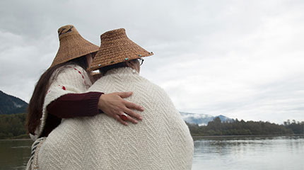 Two people wearing woven hats and blankets, standing together beside the water